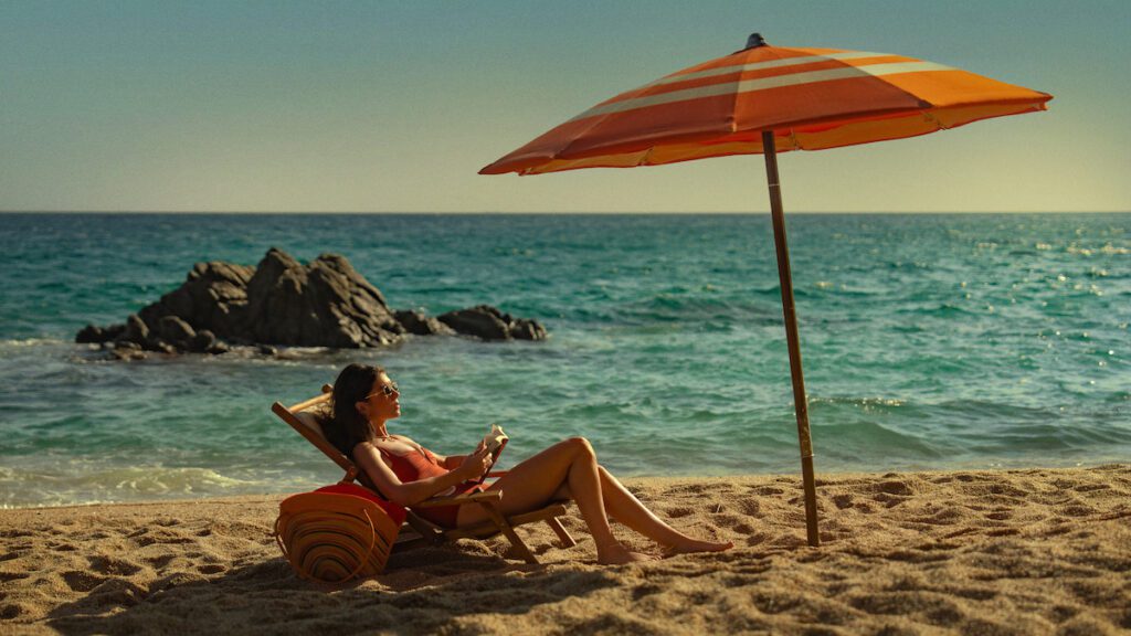 A woman relaxing on a beach lounge chair under an orange umbrella, reading a book by the ocean on a sunny day in People We Meet on Vacation