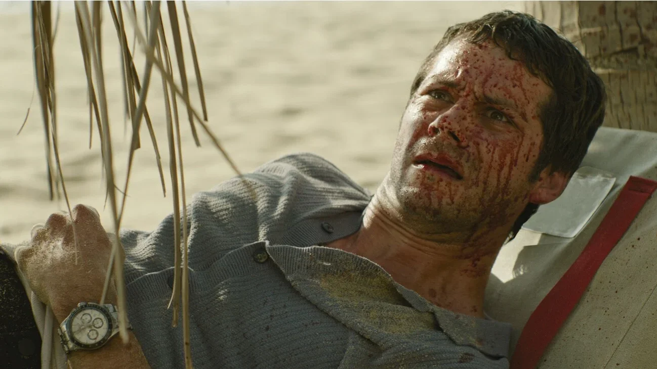 Dylan O'Brien injured and covered in blood lying on a beach after a plane crash in the movie Send Help