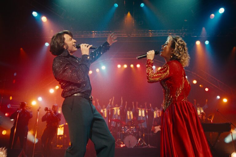 Hugh Jackman and Kate Hudson performing together on stage in “Song Sung Blue,” singing into microphones under colorful concert lights with a live band behind them.